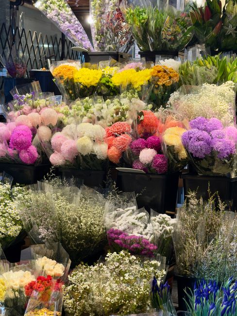 A vibrant display of fresh flowers arranged in black containers within a florist shop, featuring a variety of colours including pink, white, yellow, purple, and red. The arrangement includes different