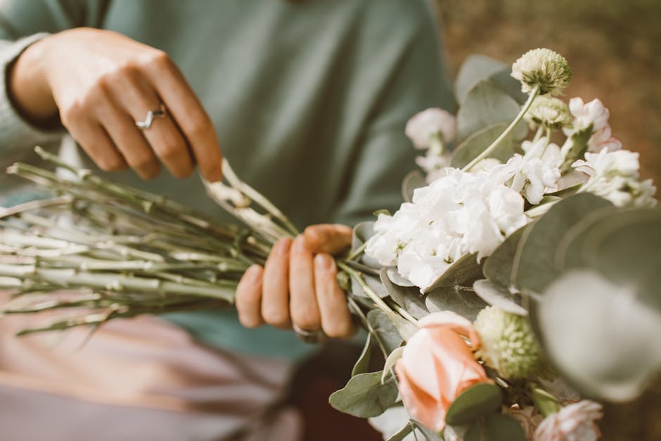 A florist's hands, adorned with silver rings, gently arrange a bouquet of fresh flowers featuring white hydrangeas, pale pink roses, and green chrysanthemums. The bouquet is wrapped in light grey tiss