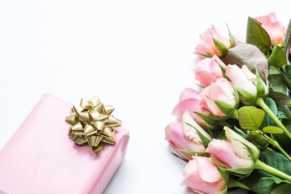 A neatly wrapped gift box in soft pink paper with a shiny gold bow sits beside a lush bouquet of pale pink and white roses with green stems and leaves. The flowers are arranged on a white surface, sho